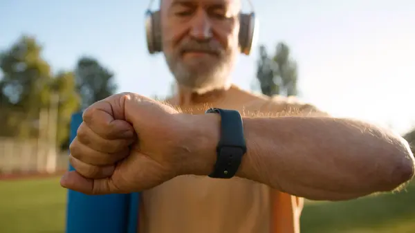 Pensioner looking at wristwatch end of training old Caucasian man leaving after sport physical activity exercise health sportsman in headphones listening music gymnastics aerobics stadium city outside