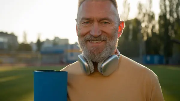 Old Caucasian man holding mat sportsman physical activity lifestyle pensioner looking around smiling sport headphones stadium city outside fitness healthcare athlete training warm-up equipment morning