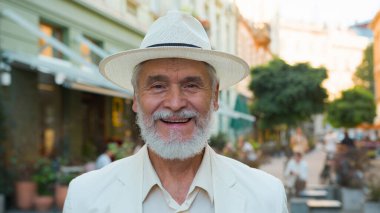 Mutlu gülümseyen yaşlı, yaşlı, yaşlı, olgun, beyaz saçlı, zarif iş adamı, turist, şehrin dışındaki tatil günlerinde kameraya olumlu bir yüz ifadesiyle bakıyor.