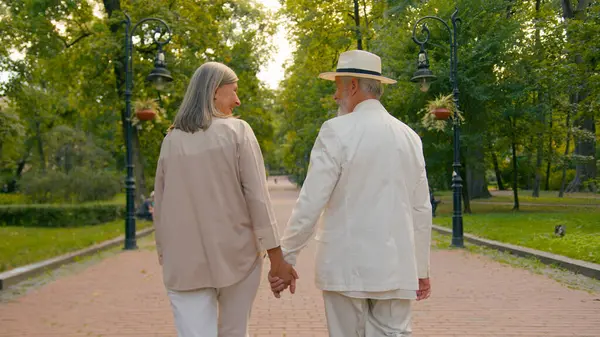 Back view happy Caucasian family old couple man woman holding hands walking together city park outside gray-haired senior husband wife enjoying walk health care retirement marriage active lifestyle