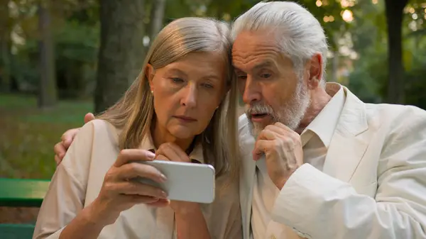 Confused worried Caucasian old senior couple retired family elderly man woman looking smartphone receiving bad news notification negative reaction sadness worry upset losing failure outside park bench