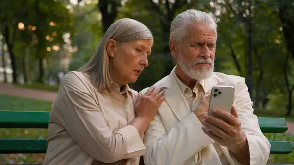 Upset puzzled nervous disappointed aged Caucasian family couple elderly man woman holding mobile phone talking discuss difficulties wireless modern gadget device problem error repair outside city park