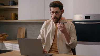 Bilgisayarla çalışan çok görevli beyaz adam ev mutfağındaki telefonla sesli mesaj kaydediyor. Mobil uygulamayla uzaktan iletişim kuran serbest çalışan erkek işadamı.