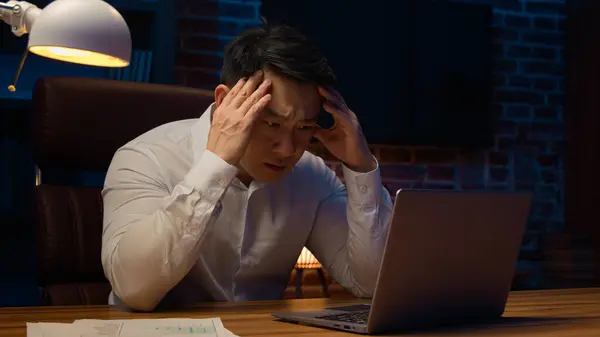 Stressed exhausted ill sad tired Asian mature man overworked Korean businessman at night office late time stress with business project problem suffer headache trouble solution computer laptop failure