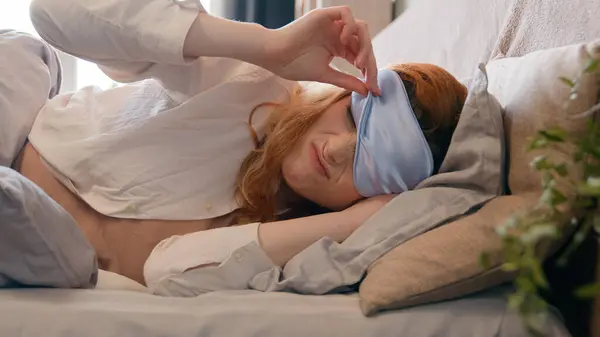 Sleeping napping lazy Caucasian girl with blindfold in bed in early morning peek out from under sleep mask open eye fall asleep again woman sleepy female angry hear sound of alarm clock waking up