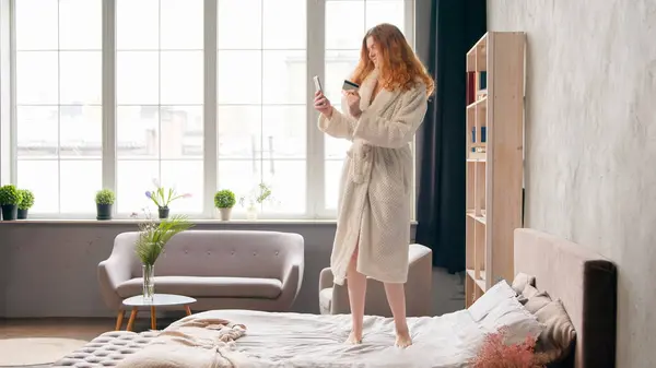 stock image Caucasian girl happy carefree woman in bathrobe shopping online with phone and credit bank card success mobile payment dancing celebrate successful buying purchase discount in morning bedroom on bed