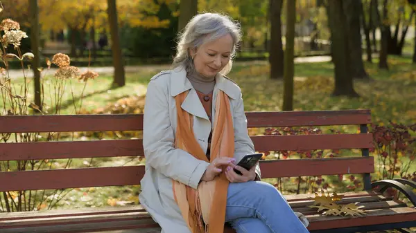 Happy grey-haired beautiful mature woman using smartphone online in autumn park Caucasian female using internet technologies outside senior lady browsing mobile phone apps looking at screen outdoors