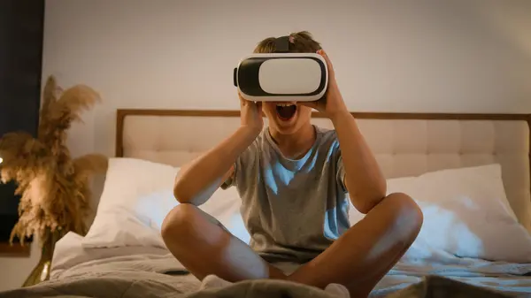 Surprised amazed Caucasian child kid boy playing video game using innovative digital VR headset on bed home play online 3d AR technology in metaverse world wearing wireless virtual reality glasses