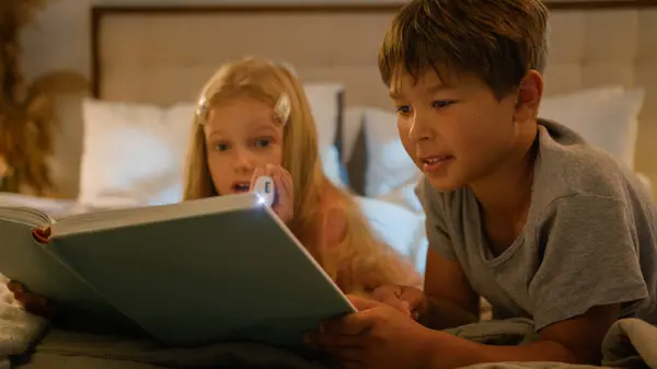 stock image Caucasian children kids boy girl older brother younger sister lying on bed at home evening reading interesting book using flashlight read fairytale story for night bedtime happy family relationships
