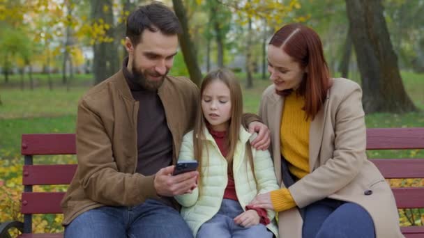一緒に幸せな男 父親の女性 母親の子供 屋外公園 ベンチパパパママ 赤ちゃん 携帯電話のスマートフォンブラウジング オンラインで話す商品を選択する 都市で話す — ストック動画
