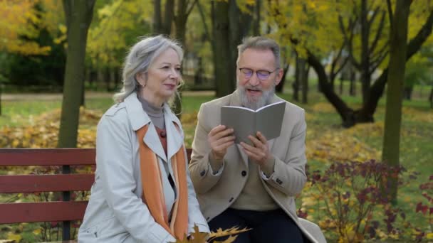 老两口老年成熟女性白种人家庭聪明的退休女性男性阅读诗歌在公园的长椅上看书丈夫微笑快乐的祖父母享受户外休闲 — 图库视频影像