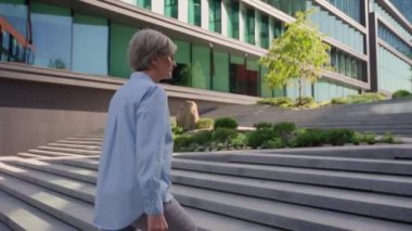 Geriye dönüp bakıldığında, kıdemli beyaz iş kadını kadın işkadını şehir dışında merdivenlerden yukarı çıkıyor mutlu orta yaşlı, memnun bayan işveren merdivenleri tırmanıyor kameraya gülümsüyor.