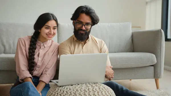 Multiracial couple Arabian man and Indian woman on floor family couple homeowners wife girlfriend and husband boyfriend watching laptop movie smart TV enjoy home leisure smiling talk video call smile