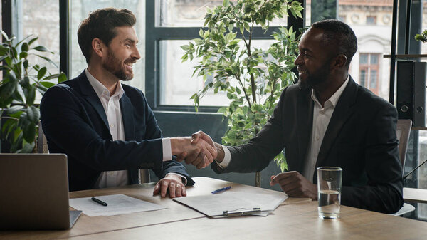 Two diverse multiracial businessmen Caucasian man and African American businessman partners men signing contract agreement document shaking hands conclude success business deal partnership in office