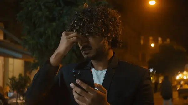 Stock image Indian Arabian Latino man holding mobile phone indignant confused angry displeased mad ethnic male cellphone user technology city outside night negative trouble problem spam furious notification lost