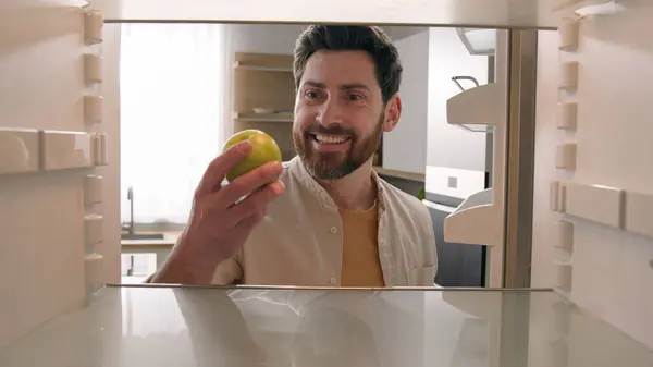 POV point of view from inside refrigerator Caucasian adult happy man at kitchen open empty fridge take last green apple bite eat fresh healthy fruit hungry guy male eating diet food delivery service