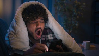 Arap yorgun esneyen adam cep telefonu kullanıyor. Uykusuzluk çekiyor. Yorgun, yorgun, uykulu, battaniye altında uykulu, sosyal medya uygulaması okuyor.