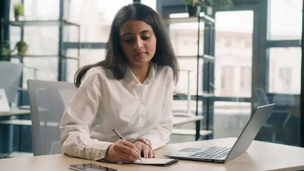 Multiracial student girl studying online serious busy businesswoman in office writing notes ideas business tasks in notebook working with laptop Indian Arabian woman manager worker at workplace work