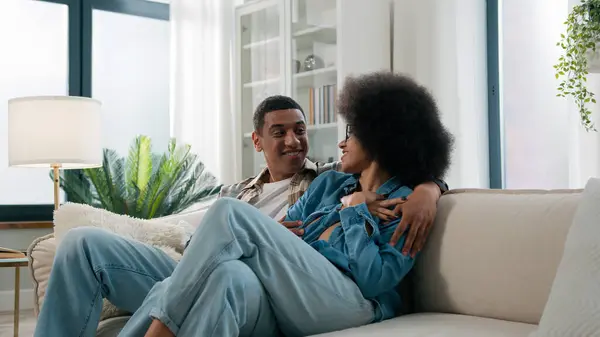 African American spouses couple man woman girl guy on sofa couch at home own apartment together talk discuss family affairs plan smiling laughing talking hugging bonding husband wife nice conversation