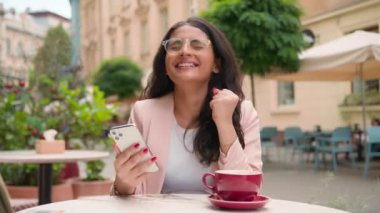 Mutlu, mutlu, gülümseyen Hintli, Arap, etnik kadın kız zaferi kutlayan bir cep telefonu mesajı aldı. Şok gülümsemesi kazanıyor. Şehir kafesinin önünde gülüyor. İnternet üzerinden.