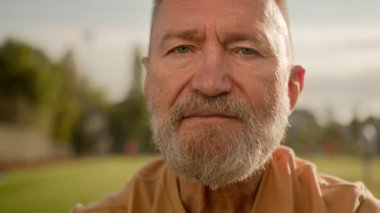 Kameraya bakan yaşlı beyaz adam ciddi yüz ifadesine bakıyor. Şehrin dışındaki sakin, göz kırpan spor stadyumu. Sakallı, buruşuk sakallı, erkek yaşam tarzı.