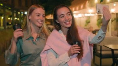 Açık havada gezen iki Avrupalı genç kadın telefon konuşmaları yapan akıllı telefon arkadaşlarıyla sohbet ederek gülüşmeler, kahkahalar, kahkahalar, akşam kahveleri vlog turizmi çevrimiçi kentsel selfie
