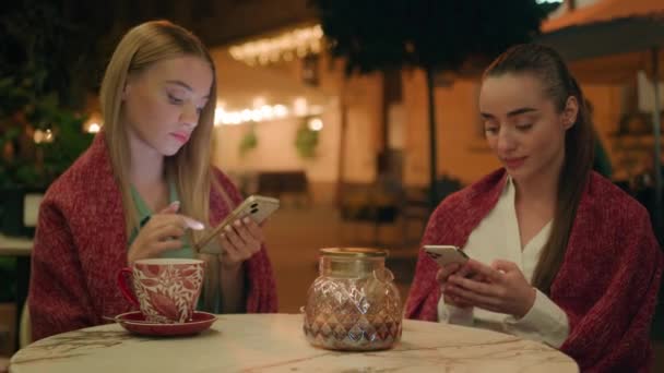 2人の若い白人女性カフェ都市外で携帯電話のスマートフォンテキストチャットウェブを使用して夜は一緒に茶の居心地の良い女の子の姉妹の友人を飲みますインターネットソーシャルメディア接続を満たす — ストック動画
