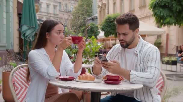 コーカサス カップル 朝食を食べる 街のカフェ 屋外で 忙しい男 携帯電話を使用して ガールフレンド 怒った腕 クロス — ストック動画