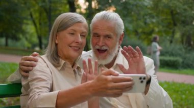 Yakın çekimde gülümseyen beyaz orta yaşlı bir aile ferdi akıllı telefonları şehir parkında dinlendirici konuşuyorlar. Mutlu büyükanneler ve büyükbabalar çevrimiçi video: