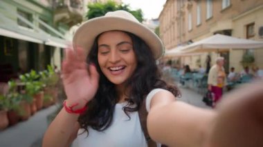 Genç, mutlu, gülümseyen Hintli Arap bayan öğrenci kız öğrenci sosyal medya vlogcusu elinde akıllı telefon kamerasıyla kamera çekimleriyle sokak dışında sanal telefon görüşmesi yapıyor.