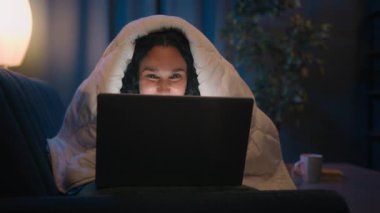 İspanyol gülümseyen mutlu Arap kadın geceleri evde bilgisayar tarama laptopu kullanıyor. Battaniye oyunu bağımlılığı son teslim tarihi. Karanlık akşam çalışma saatlerinde internet alışverişi yapıyor.