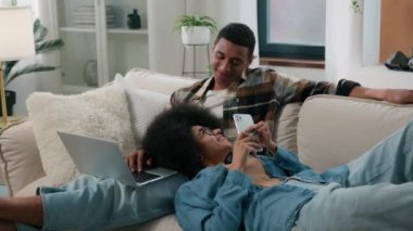 Dizüstü bilgisayarlı Afro-Amerikalı çift, kanepede oturmuş akıllı telefonlarıyla sohbet ediyor, cep telefonlarını karıştırıyor erkek dizlerinin üzerinde yatıyor, kız arkadaş, alet bağımlısı, internet bağımlısı, gülümseyen bir aile.