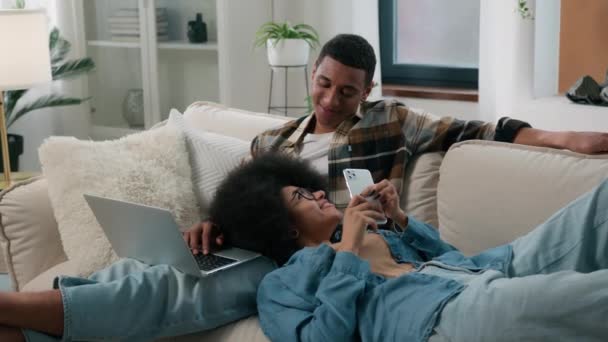 アフリカ系アメリカ人のカップル男 ソファの家に座ってノートパソコンの女性チャット スマートフォンブラウジング 男性の膝に横たわる携帯電話ガールガジェット中毒インターネット中毒笑顔家族 — ストック動画