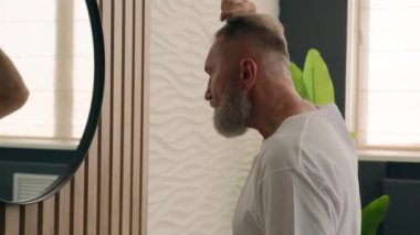 Beyaz sakallı yaşlı adam otelin banyo fırçasında aynaya bakıyor saçlarını tarıyor olgun orta yaşlı emekli erkek saç fırçasıyla saçlarını tarıyor.