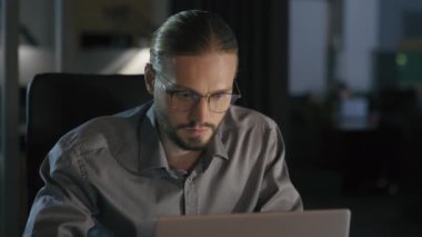 Gece fazla mesai yapan işadamı yöneticisi karanlık ofis bilgisayarı ile çalışıyor dişlek, mutlu iş adamı lider işveren masa başında bilgisayarla kameraya bakıyor.