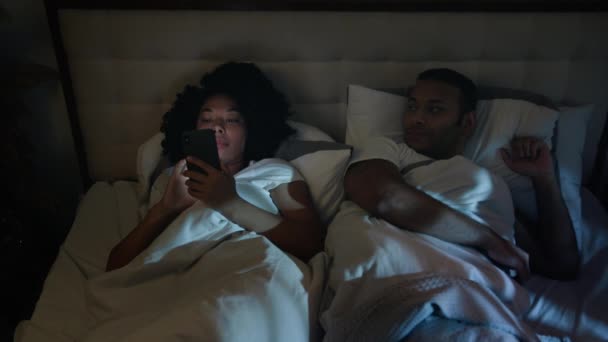 アフリカ系アメリカ人のガジェット中毒ソーシャルメディア携帯電話中毒女性 ベッドの夜にスマートフォンを使用して悲しい怒り不満の夫を分離した妻 睡眠を試みる 時間遅くの注意が必要 — ストック動画