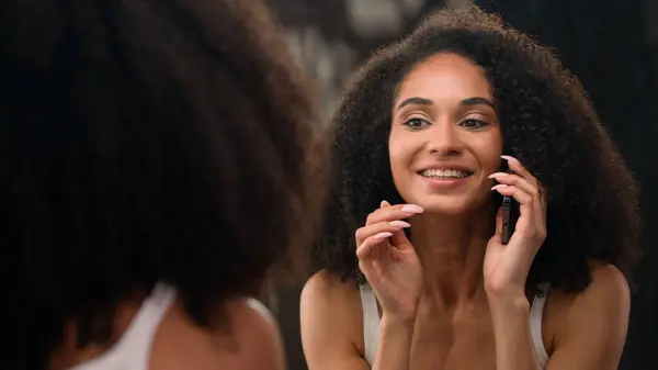 Beautiful African American woman happy smiling girl looking at mirror reflection touch face make up preparing skin care moisturize with cream skincare procedure talking mobile phone call smartphone