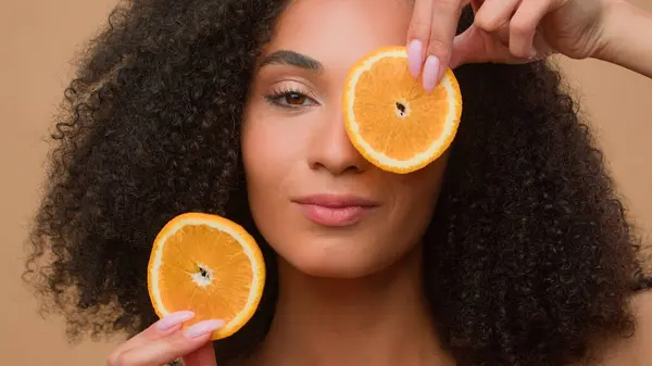 ベージュの背景アフリカ系アメリカ人女性ビタミン栄養化粧品スキンケア保湿オイル血清フェイシャルクリームの2つのオレンジ柑橘類のスライスを保持する民族モデルを閉じる — ストック写真