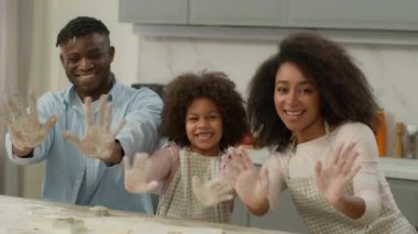 Afro-Amerikan mutlu aile etnik ebeveynleri ve küçük kız çocukları mutfakta hamuru pişirirken gülümserken kameraya bakıp kirli avuç içi un lekelerini gösteriyorlar.