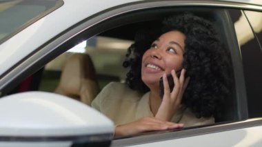Neşeli müşteri Afrikalı Amerikalı kadın zengin iş kadını müşteri alıcı etnik kız heyecan içinde yeni otomobil modern lüks araba içinde oturuyor cep telefonu konuşarak mutlu bir şekilde galeriden araba satın alıyor.