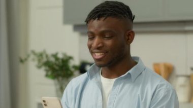 Mutlu gülümseyen Afro-Amerikan etnik erkek kullanıcı kaygısız etnik erkek gülümsemesi akıllı telefon kullanarak sosyal medya alışverişi ev mutfağında online kaydırma aparatı
