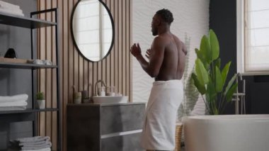 Neşeli Afrikalı, seksi, etnik erkek hipster, kalçasında havluyla dans ediyor, dans ediyor, banyoda müzik dinliyor, hijyenik, neşeli Amerikalı erkek kameraya parmak işareti yaparak dans ediyor, gülümsüyor.