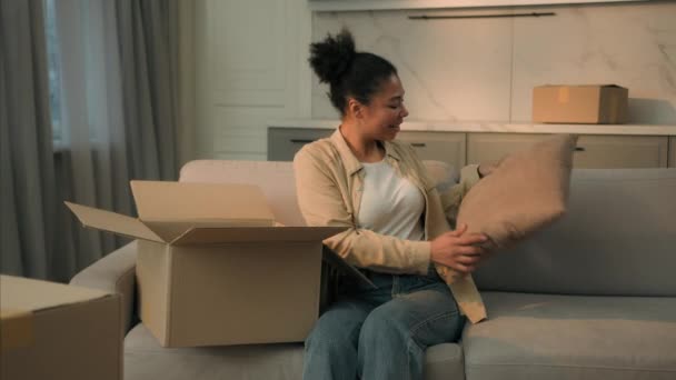 アフリカ系アメリカ人少女笑顔バイヤーアンパッキング段ボール箱購入クッション枕ソファ幸せな女性ホームオーナーが新しいアパート移転日に移動する日パックオープン小包配達オンラインショッピング — ストック動画