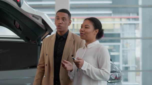 アフリカ系アメリカ人女性セールスウーマンディーラーコンサルティングクライアントは 自動車オートオープニングクロスオーバートランクの中を見るショールームの顧客の車の特徴を説明する現代自動車を選択する男性を助けます — ストック動画