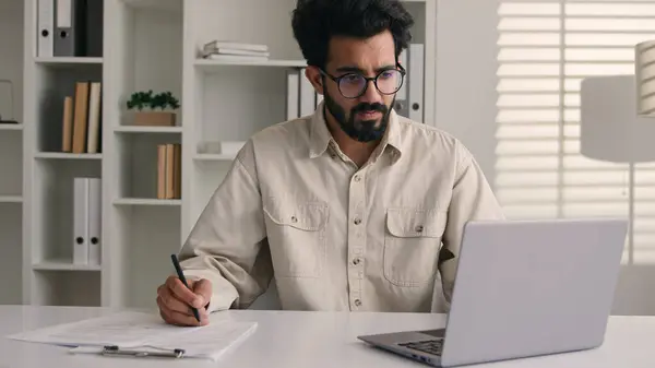 焦点人物阿拉伯裔印度裔商人男性雇员经理写便条业务任务数据文书工作签署文件在预算文件上使用手提电脑办公 — 图库照片