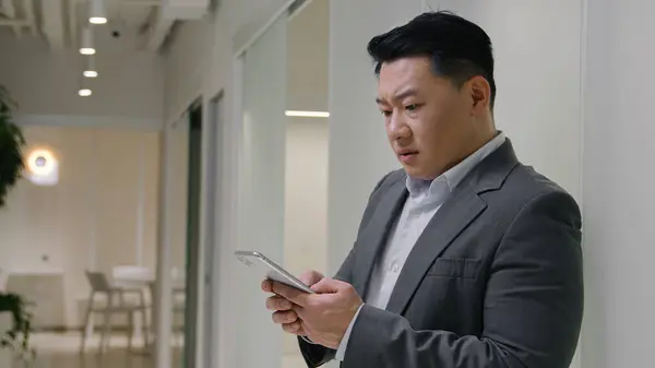 Asian man in corporate office using mobile phone upset thoughtful chinese korean middle-aged businessman texting messages online business social networking receive bad news lost think with smartphone