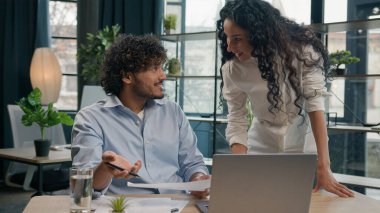 Hispanic woman leader mentor business coach help Arabian man intern with corporate laptop work at office businesswoman teach businessman explain financial paperwork new employee adaptation assistance