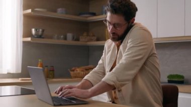 Modern mutfakta dizüstü bilgisayarı olan serbest çalışan bir erkek işadamı evden uzak bir yerde cep telefonuyla projeyi tartışıyor. 40 'lı yılların erkek ev sahibi akıllı telefon tarayıcı bilgisayar çoklu görev