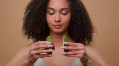 Afro-Amerikan güzel, gülümseyen kadın, kıvırcık saçlı, sağlıklı cilt bakımlı, stüdyo bej arka planda iki yarım avokado beslenme diyeti, organik vitamin kozmetik.