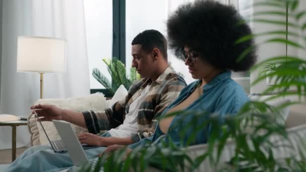 忙着工作的自由职业者非洲裔美国男人和女伴在家里沙发上浏览笔记本电脑Pc设备在线互联网自由职业者远程工作成瘾玩电子游戏应用程序 — 图库视频影像